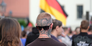 In Köthen steht ein Mann mit tätowiertem Hinterkopf vor einer Menschenmenge über der eine Deutschlandflagge weht