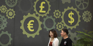 Menschen gehen an einer Wand vorbei, welche die Symbole für den Yen, Euro, Dollar und britischen Pound zeigt