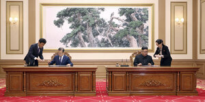 Zwei Männer, Moon Jae In und Kim Jong Un, sitzen an zwei unterschiedlichen Tischen, die nebeneinander stehen. Sie unterschreiben etwas