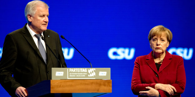 Seehofer spricht am Podium, Merkel steht daneben und zieht ein Gesicht