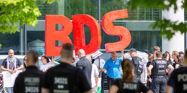 AkstistInnen halten riesige Buchstaben "BDS" hoch, davor stehen PolizistInnen
