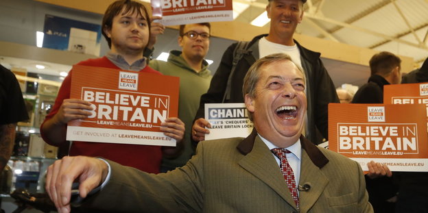 Nigel Farage lacht, hinter ihm stehen Männer mit Plakaten