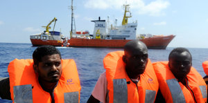 Drei Männer tragen orangefarbene Schwimmfesten, dahinter sieht man ein Schiff auf dem Meer