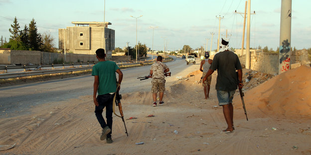 Auf einer Straße laufen drei zivil gekleidete Männer mit Waffen in der Hand