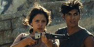 Ava und Juan stehen nebeneinander und sie hält ein Gewehr in der Hand.