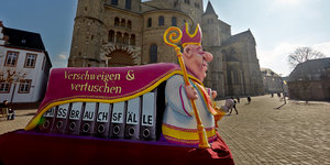 Ein Karnevalswagen zeigt einen Bischof, der seinen Mantel über die Akten mit Missbrauchsfällen wirft.