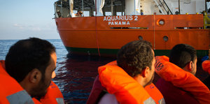Drei Männer in orangefarbenen Schwimmwesten blicken von einem Boot auf ein Schiff