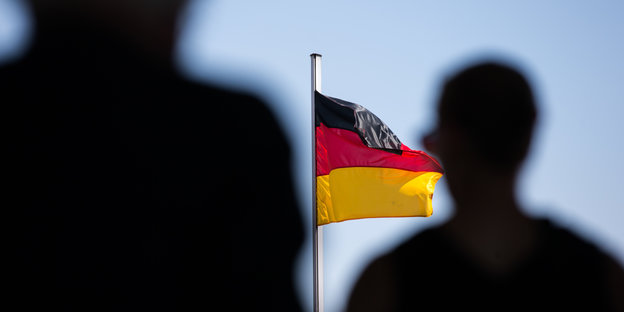 Eine Deutschlandfahne weht an einem Fahnenmast, davor stehen zwei Menschen.