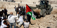 Mehrere Menschen demonstrieren vor einem Bulldozer, ein Mann hält eine palästinensische Flagge.