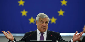 Tajani sitzt von einer EU-Flagge und macht eine fragende Geste