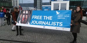 Menschen stehen mit einem Plakat mit der Aufschriff "Lasst die Journalisten frei" vor dem Hauptbahnhof