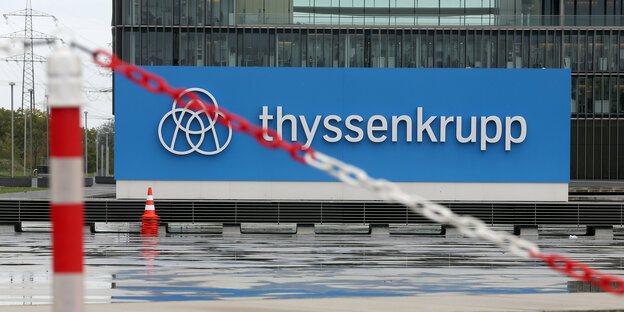 Eine rot-weiße Baustellen-Absperrkette hängt vor dem Firmenlogo von ThyssenKrupp in Essen.