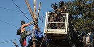 Eine halbnackte Frau hängt an Baumstämmen, daneben Polizistinnen in Kampfmontur auf einer Art Podest