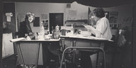 Foto eines Schwarz-Weiß-Fotos, auf dem zwei Leute an unaufgeräumten Schreibtischen sitzen
