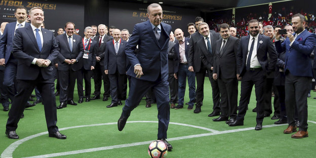 Recep Tayyip Erdoğan schießt einen Fußball, sehr viele Männer in Anzügen schauen ihm dabei zu