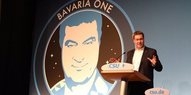Markus Söder steht an einem Pult, dahinter ein Logo mit Söders Gesicht und Schriftzug "Bavaria One"