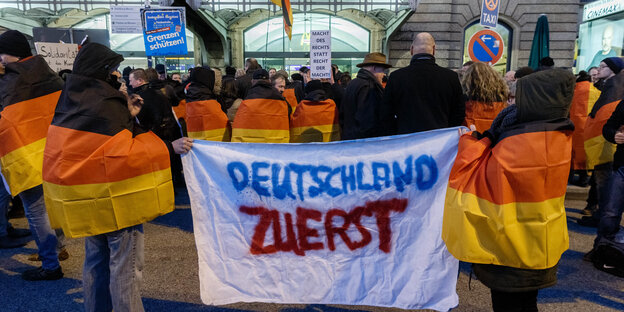 Menschen in schwarz-rot-gelbe Fahnen gehüllt, halten Transparent hoch. Darauf steht: "Deutschland zuerst"