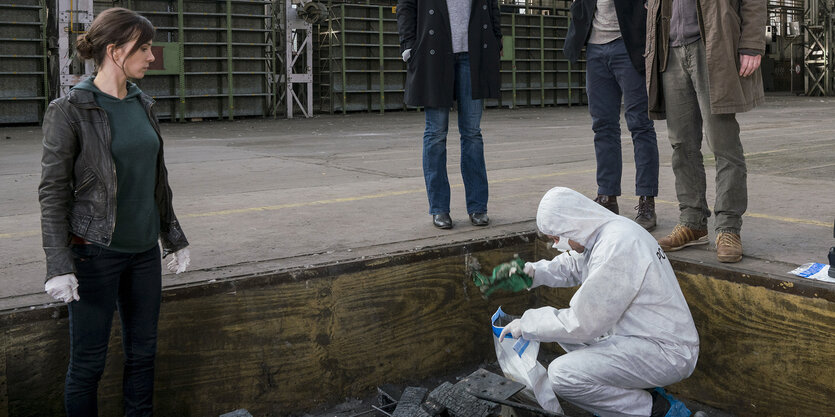 Ein Spurensicherer in weißem Ganzkörperanzug hockt an einem Tatort, daneben eine Ermittlerin