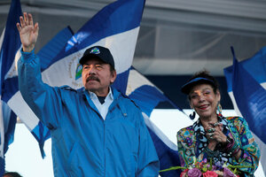 Ein Mann hebt seinen Arm zum Gruß, neben ihm applaudiert eine Frau, im Hintergrund blau-weiße Flaggen