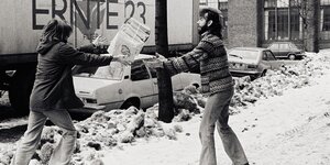 Eine Frau wirft einem Mann auf verschneiter Straße einen Stapel Zeitungen zu