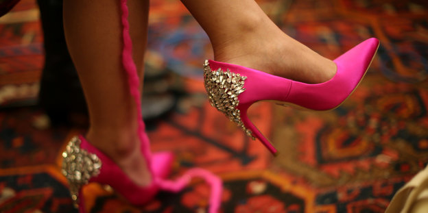 Pinke hochhackige Schuhe mit silberner Verzierung. Daneben hängt ein pinker Wollfaden herunter. Die Frau, die die Schuhe trägt strickt einen sogenannten „Pussyhat“, eine pinke Mütze