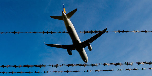 Ein Flugzeug fliegt vor blauem Himmel, dahinter Stacheldraht