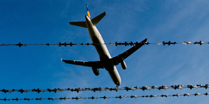 Ein Flugzeug fliegt vor blauem Himmel, dahinter Stacheldraht