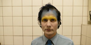 Ein Mann mit blauer und goldener Farbe auf der Stirn steht in einem Badezimmer