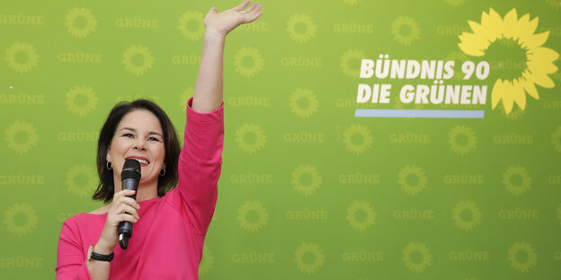 Annalena Baerbock winkt und spricht in ein Mikro, hinter ihr das Logo der Grünen