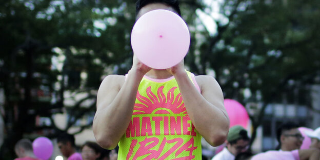 Ein Mensch in einem gelb-pinken Tanktop bläst einen rosa Luftballon auf, sein Gesicht wird dadurch verdeckt
