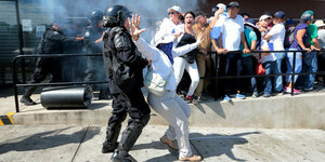 Ein Polizist in schwarzer Schutzmontur zerrt an einer weiß gekleideten Demonstrantin, im Hintergrund schreien Menschen