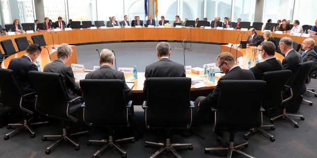 Männer in überwiegend schwarzen Anzügen und ein paar Frauen sitzen um einen großen runden Tisch