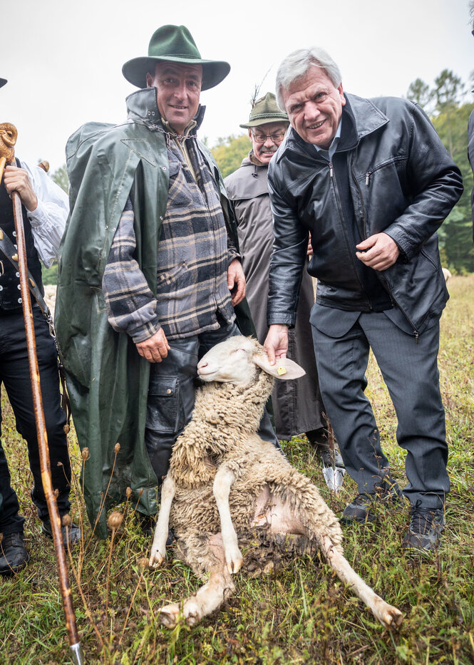 Zwei Männer stehen auf einer Weide, zwischen ihnen ein Schaf. Der linke Mann ist Schäfer und trägt einen grünen Hut und einen grünen Mantel. Der rechte Mann ist der hessische Ministerpräsident Volker Bouffier.