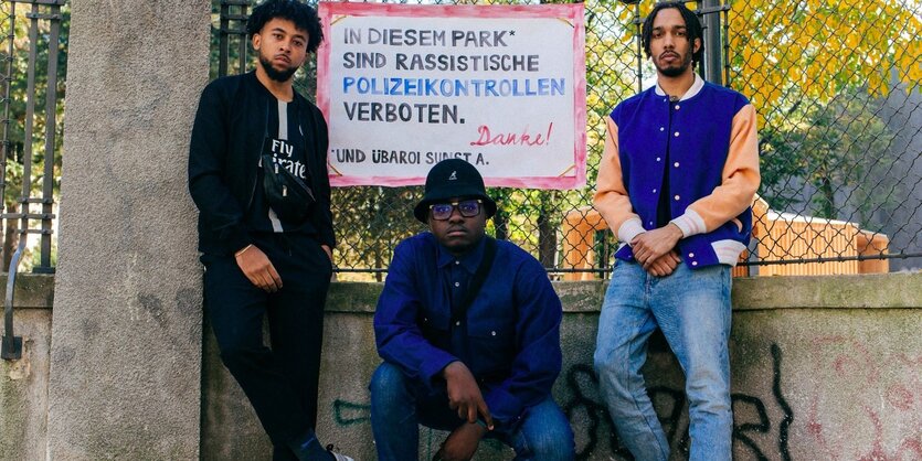 Drei junge Männer dunkler Hautfarbe vor einem Plakat vor einem Park. Auf dem Plakat steht geschrieben: "In diesem Park sind rassistische Polizeikontrollen verboten. Danke!"