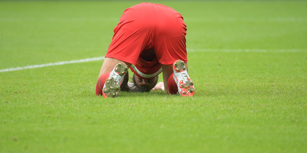 Ein Mann kniet auf einem Fußballfeld hat den Kopf auf den Rasen gelegt und den Hintern dem Betrachter entgegengestreckt