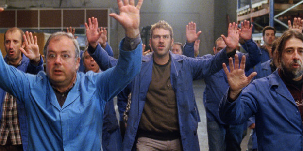 Mehrere Männer in blauen Kitteln stehen in einer Halle und halten die Hände hoch