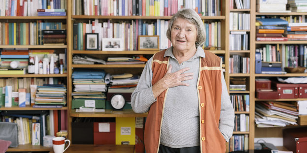 Eine ältere Frau steht vor einem Bücherregal und hält sich eine Hand vor die Brust