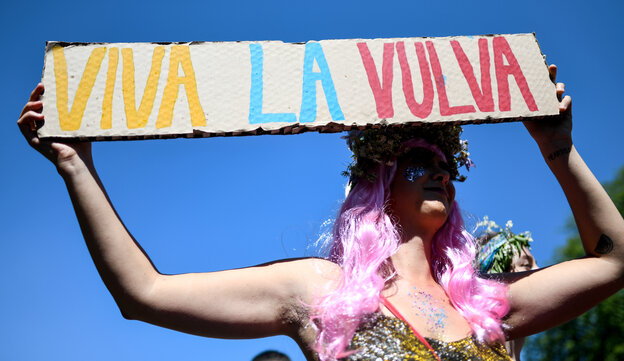 ein Mann mit rosa Perrücke in einem Kleid hält ein Plakat mit der Aufschrift "Viva La Vulva" hoch