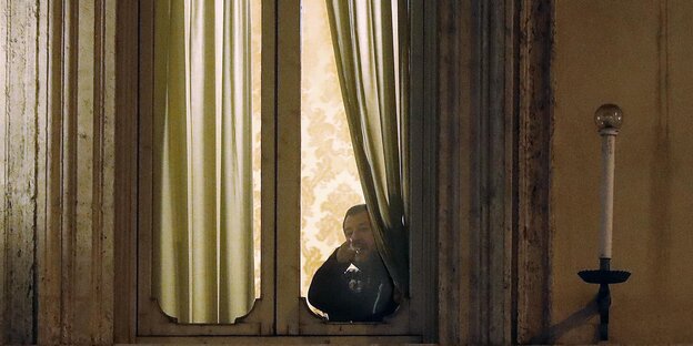 Matteo Salvini steht, halb von einem Vorhang verdeckt, an einem hell erleuchteten Fenster und guckt nach draußen