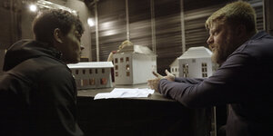 Zwei Männer sitzen an einem Tisch, der Regisseur erklärt dem Schauspieler etwas. Hinter ihnen steht das Bühnenmodell, kleine Häuser aus Pappe