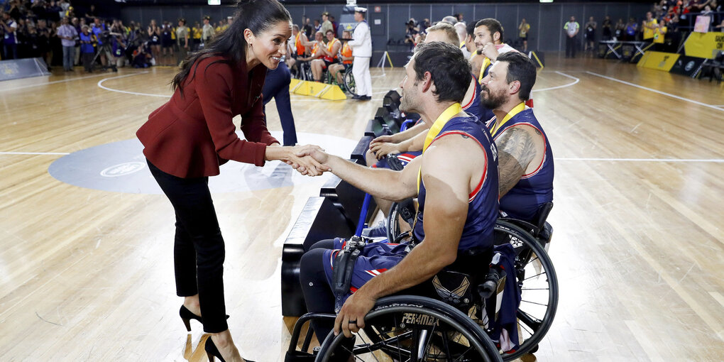 Eine junge Frau, Meghan, Herzogin von Sussex, reicht einem Mann, der in einem Rollstuhl sitzt, die Hand