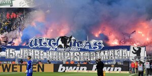 Feuerwerkskörper in dem Fanblock des Hertha BSC brennen und davor auf dem Fußballfeld stehen Spieler