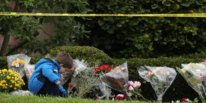 Eine Junge legt Blumen an eine Gedenkstelle, an der schon andere Blumen liegen, über ihm ein gelbes Polizei-Absperr-Band