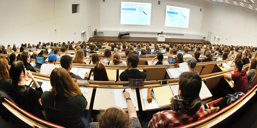 Männer und Frauen sitzen in einem Hörsaal. Sie sind von hinten zu sehen