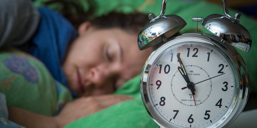 Eine Frau schläft im Bett, neben ihr auf dem Nachttischränkchen steht ein Wecker.