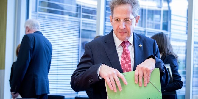 Hans Georg Maaßen packt Akten in seine tasche, im Hintergrund tritt Horst Seehofer aus dem Raum Treppengitter und hat beide Ellenbogen auf die Knie gestützt