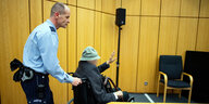 Ein Justizbeamter schiebt einen Mann im Rollstuhl in den Gerichtssaal. Der trägt einen dunkelgrauen Mantel und einen grauen Schlapphut, sein Gesicht ist verpixelt