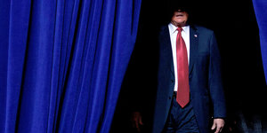 Trump tritt hinter einem blauen Vorhang hervor, das Scheinwerferlicht beleuchtet von seinem Gesicht nur den Mund