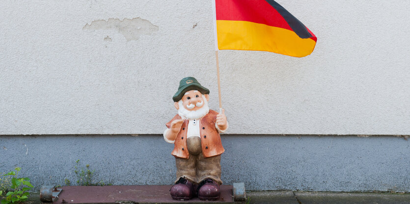 Ein Gartenzwerg steht vor einer Hauswand, in der Hand hält er eine Deutschlandflagge