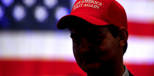 Ein Mann steht vor einer amerikanischen Flagge. Auf seinem Kopf trägt er eine Cap mit dem Spruch: Make America great again
